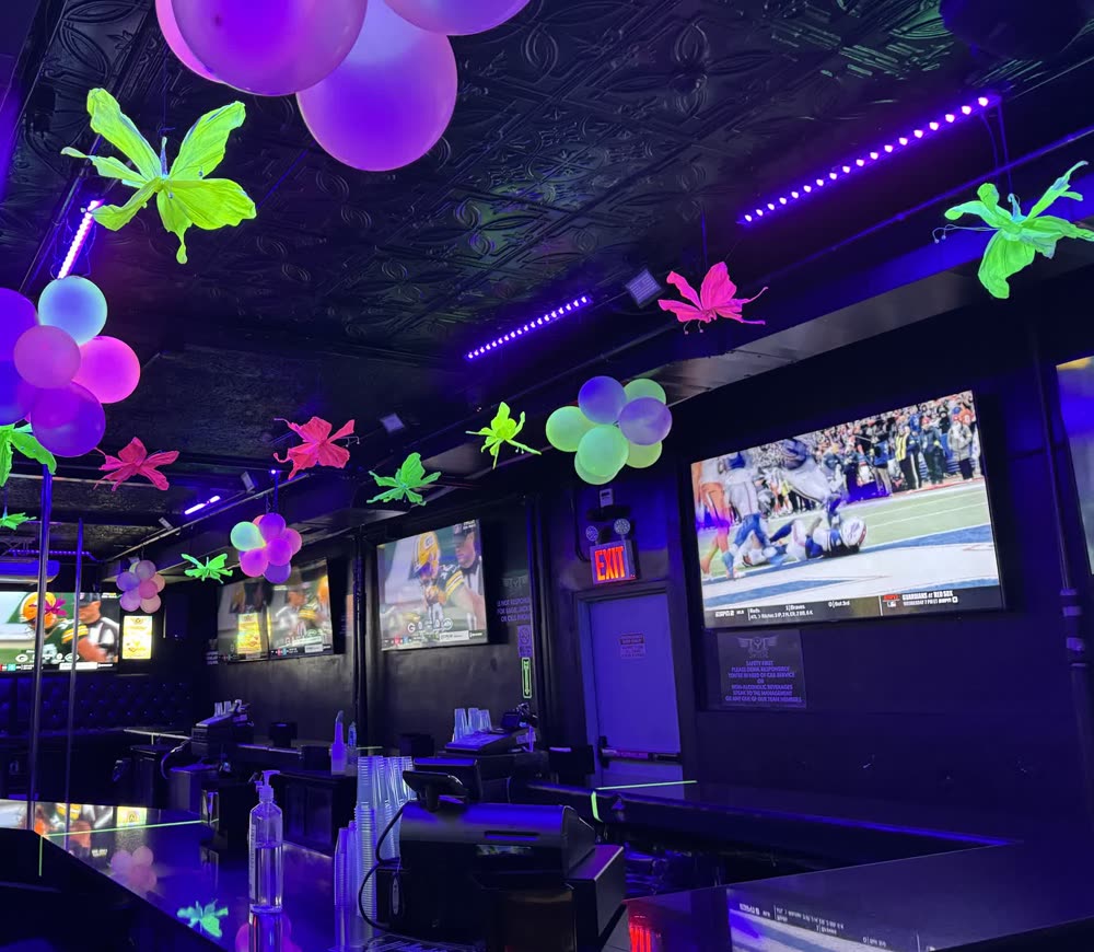 La barra de Sweets of NY decorada con globos y mariposas, pantallas grandes con deporte sobre el mostrador y luz rosa y dorada en toda la sala.
